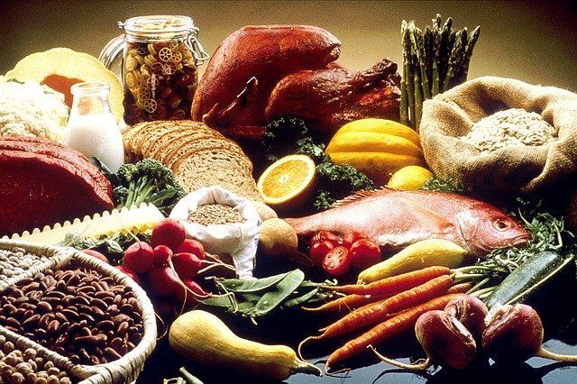 Assorted fresh foods, including meat, vegetables, beans, bread, and herbs, arranged on a table.