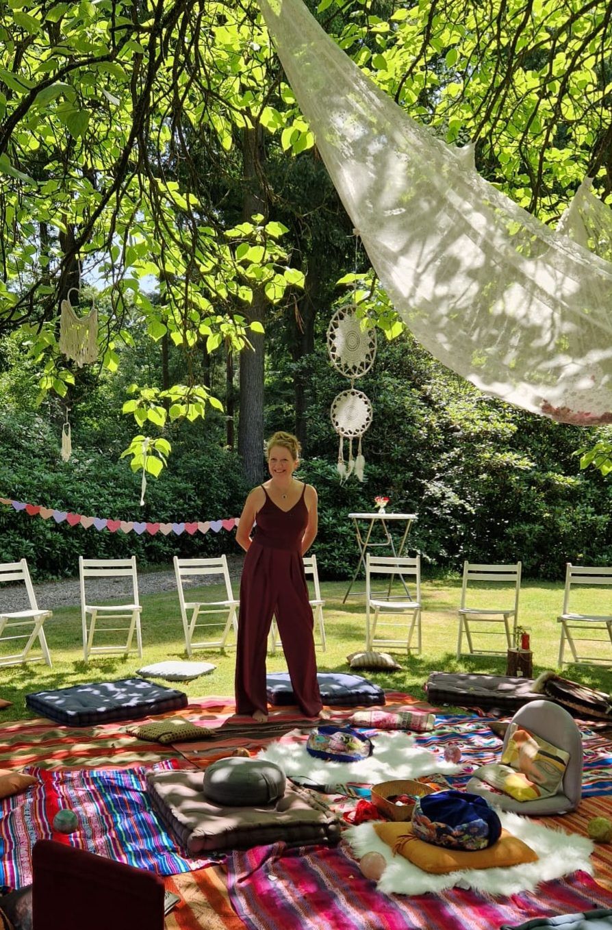 Een vrouw in een rode jumpsuit staat in een meditatiekring in de buitenlucht; kussens, dromenvangers en witte stoelen staan onder de bomen.