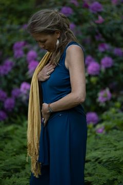 Een persoon in een blauwe jurk en gouden sjaal kijkt neer in een tuin met paarse bloemen.