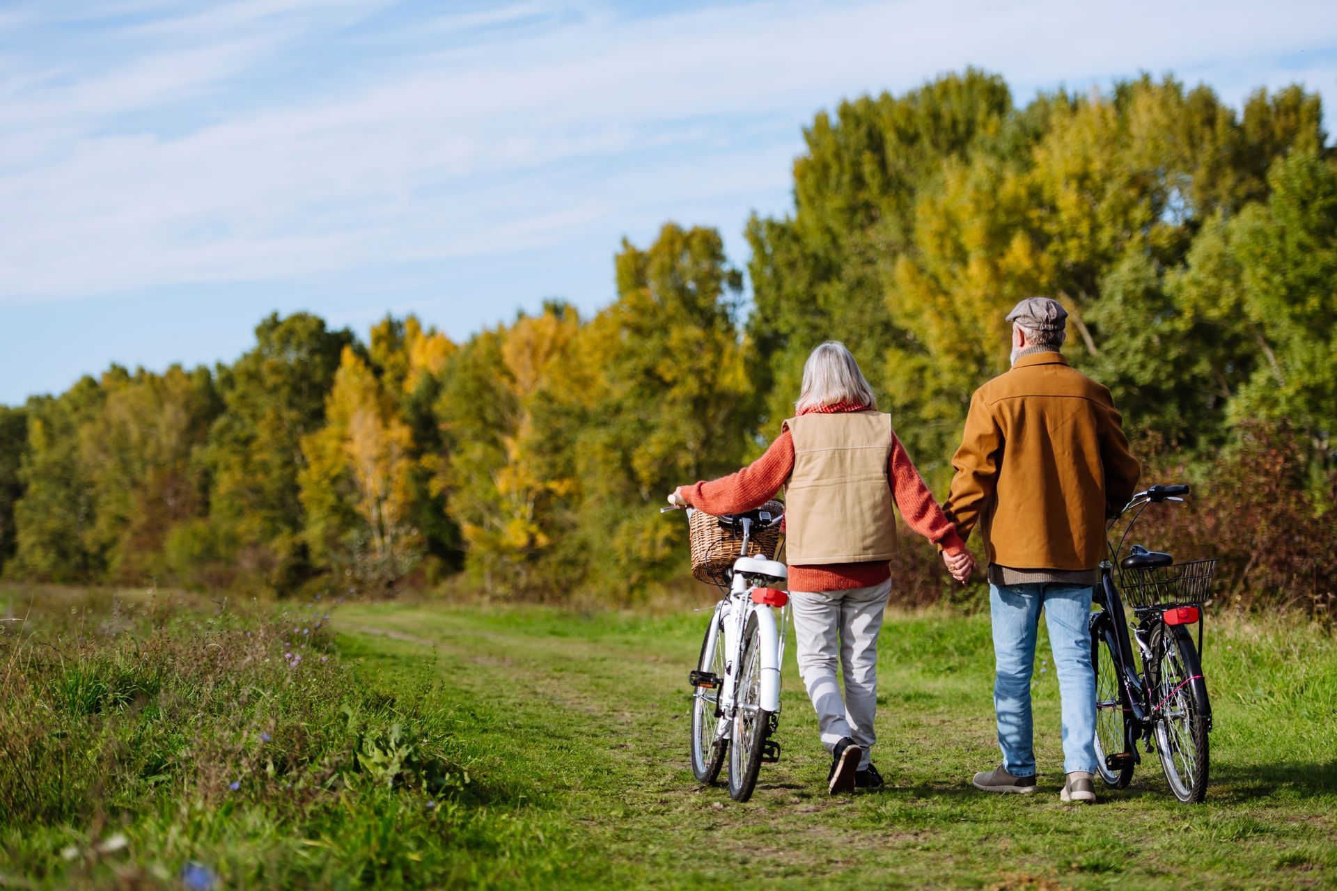 Two people with bicycles on a grassy path beside trees under a blue sky