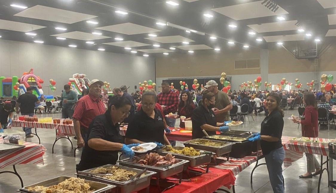 Volunteers serving food from a buffet line at a large indoor community event with tables and festive decorations.