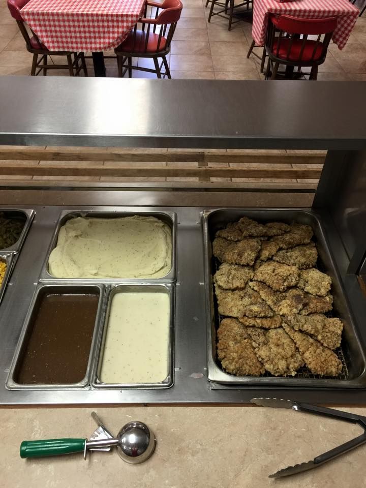 A buffet serving table with mashed potatoes, brown and white gravy, and fried meat, with a scoop and tongs on the counter.