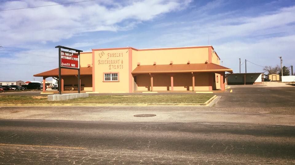 A tan commercial building with a covered porch and a prominent roadside sign under a blue sky.