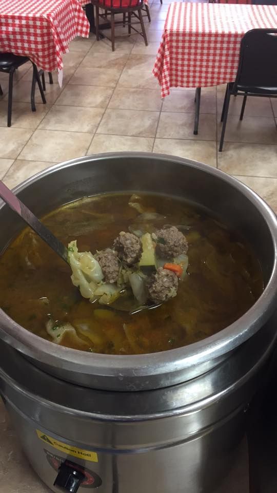 A metal ladle lifting meatballs and vegetables from a large silver soup warmer in a dining area with checkered tablecloths.