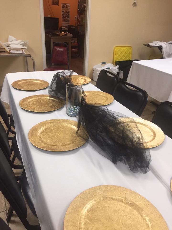 A table set with white linens, gold charger plates, and black tulle centerpieces in a dining hall.