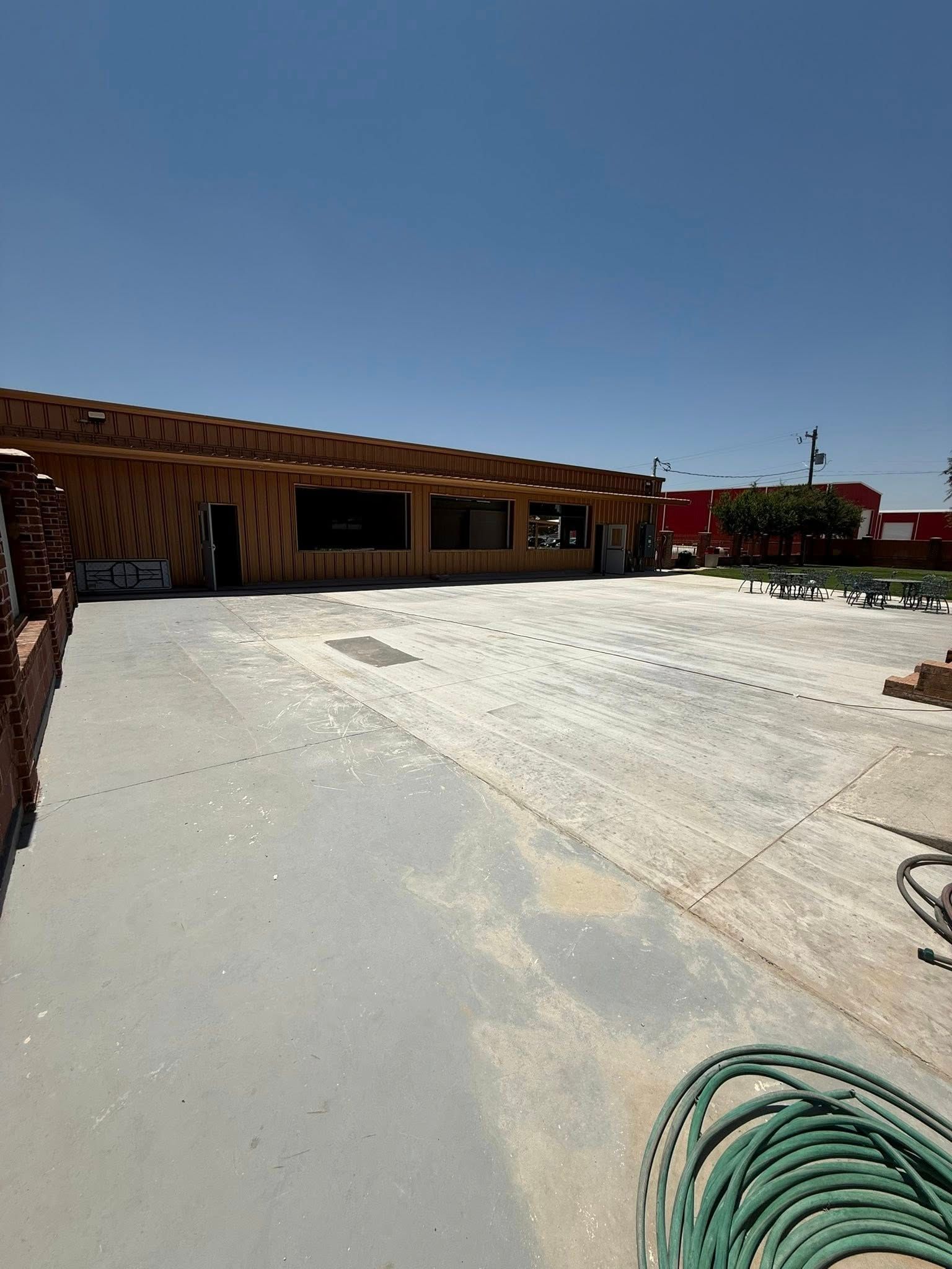 An exterior view of a low, brown building with a shaded walkway and a wide, dusty concrete patio under a clear blue sky.
