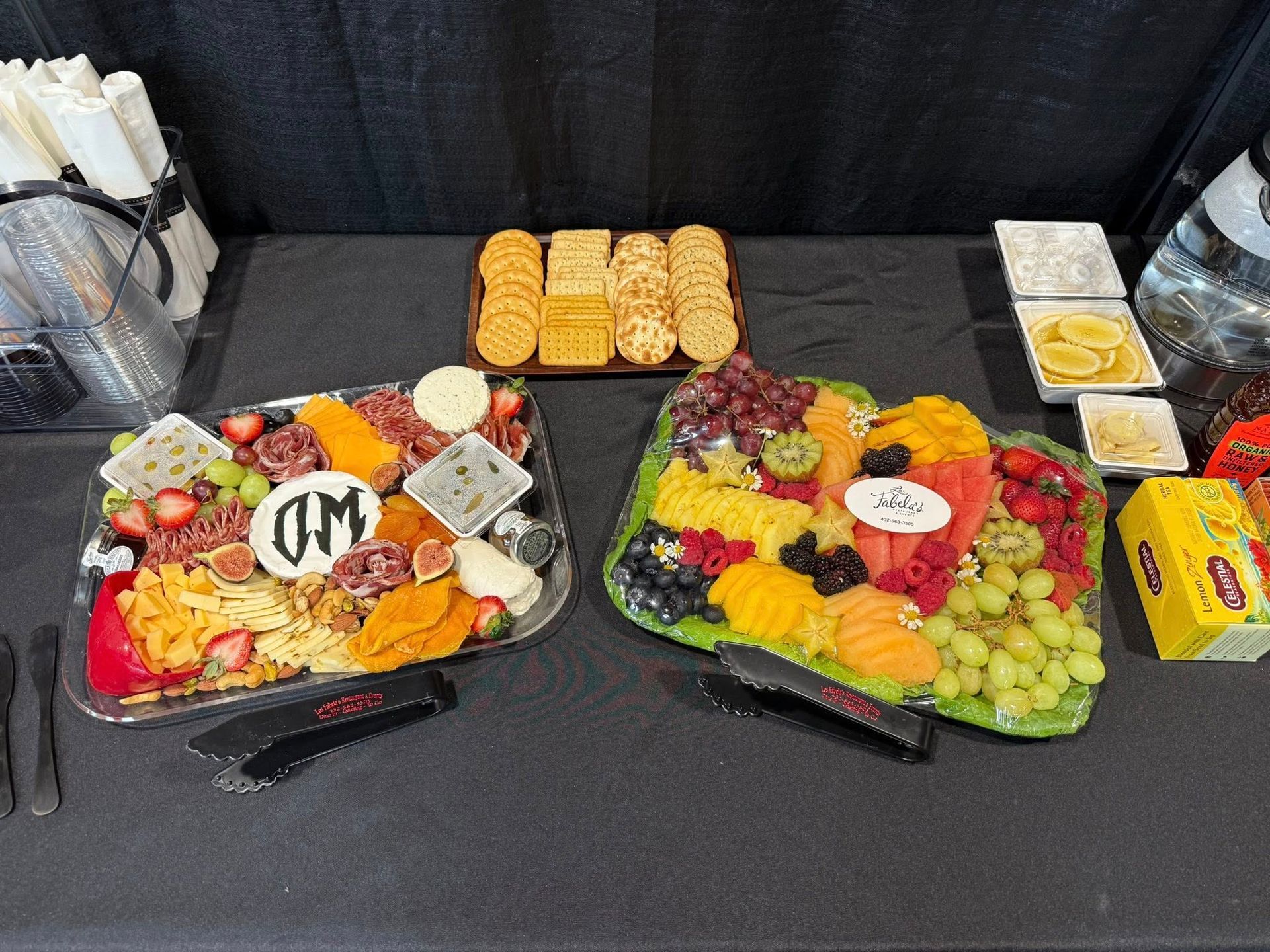 Two food platters with fruit, cheese, and meats, a cracker tray, and small bowls of dips on a black table cloth.