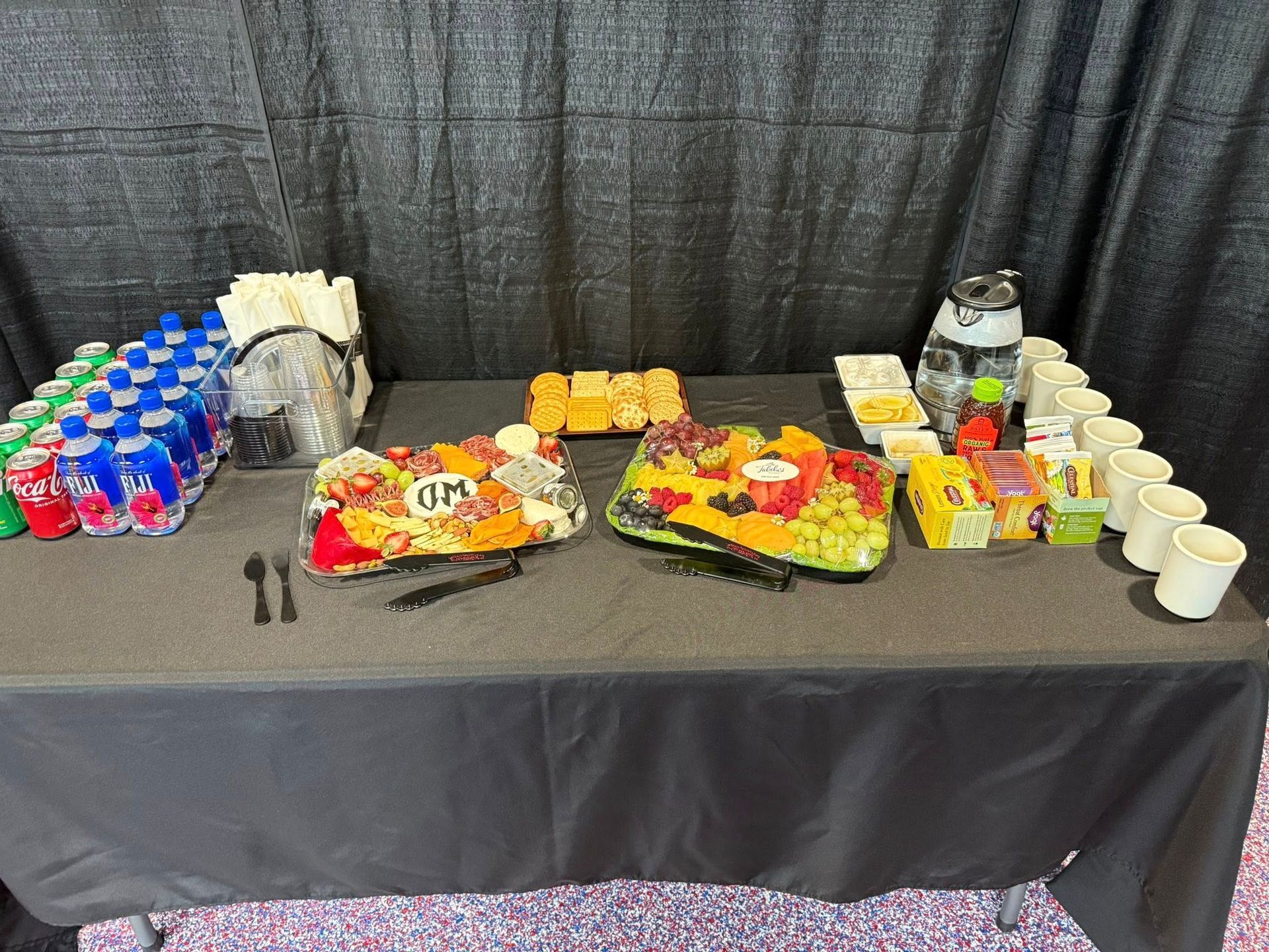 A catering table featuring two fruit and cheese platters, bottles of water, disposable cups, and small snacks.