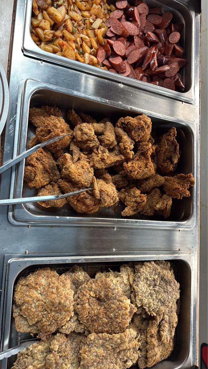 A cafeteria serving line with three metal pans containing fried chicken, sausage slices, potatoes, and fried patties.