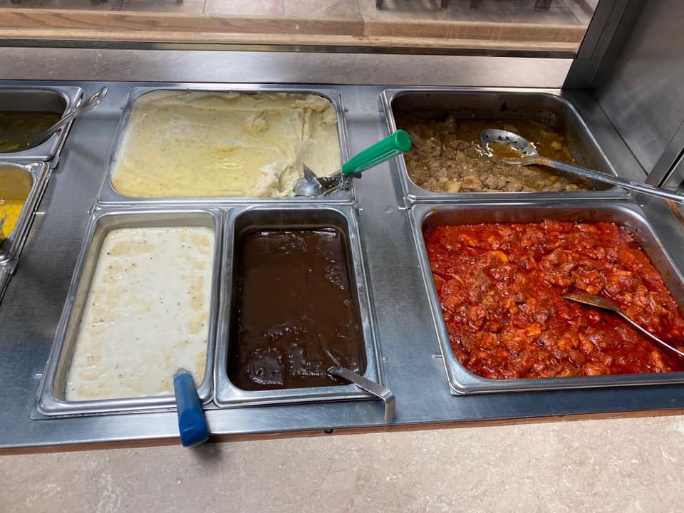 A cafeteria buffet serving line with trays containing mashed potatoes, brown gravy, white sauce, and red sauced food.