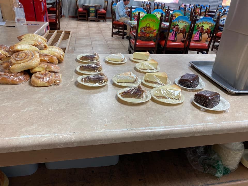 A variety of pastries and dessert slices on small plates arranged on a restaurant counter near empty dining tables.