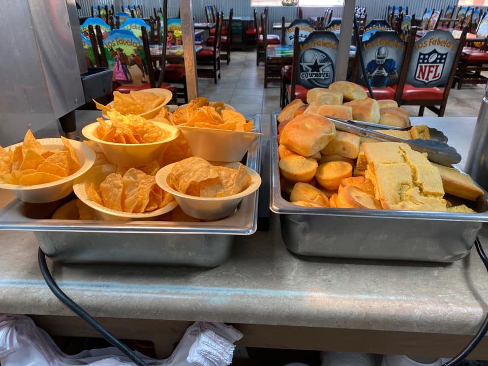 Two metal catering trays on a counter containing bowls of tortilla chips, dinner rolls, and slices of cornbread.