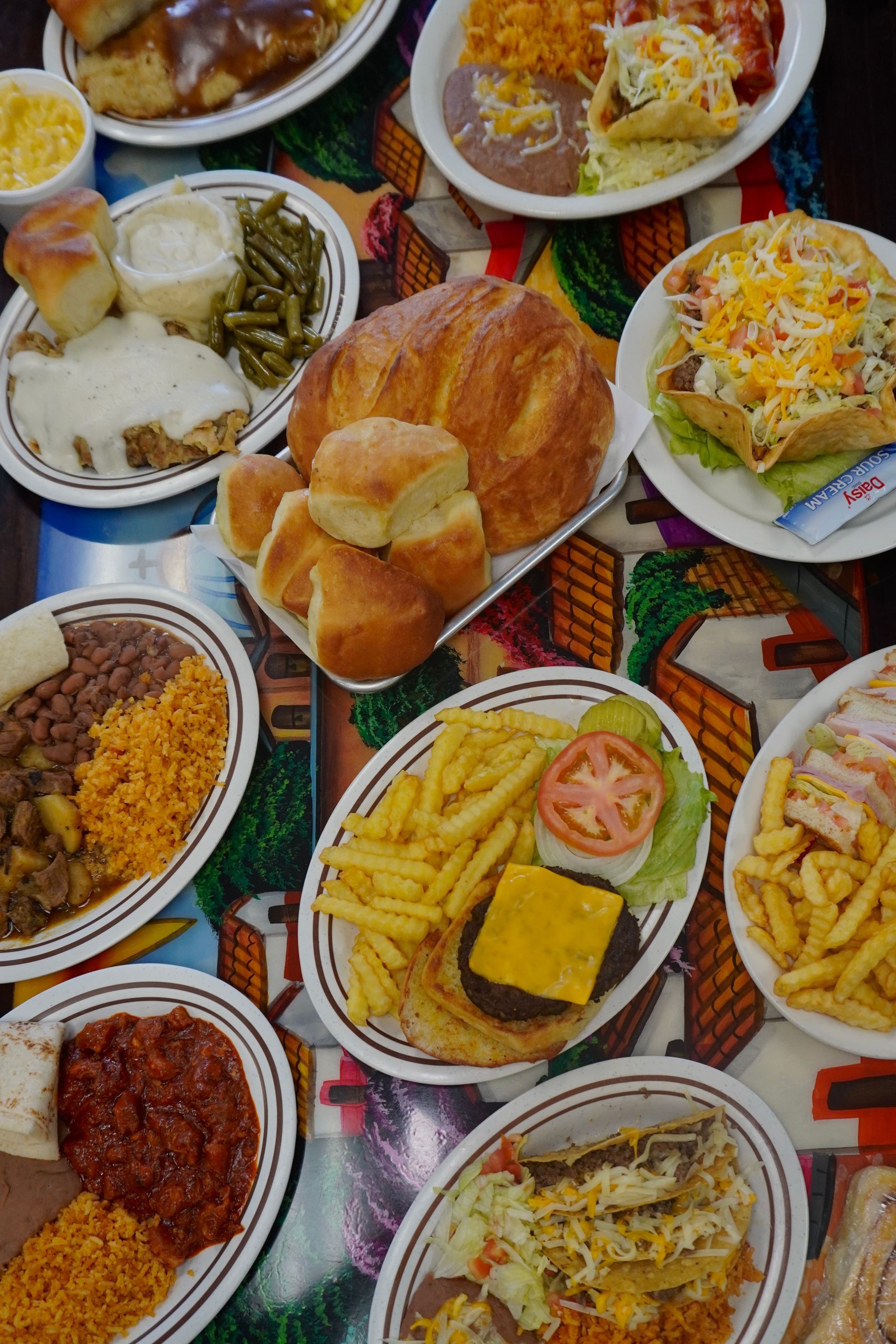 Three metal cafeteria trays containing fried chicken, sliced sausage, potatoes, and breaded meat patties.