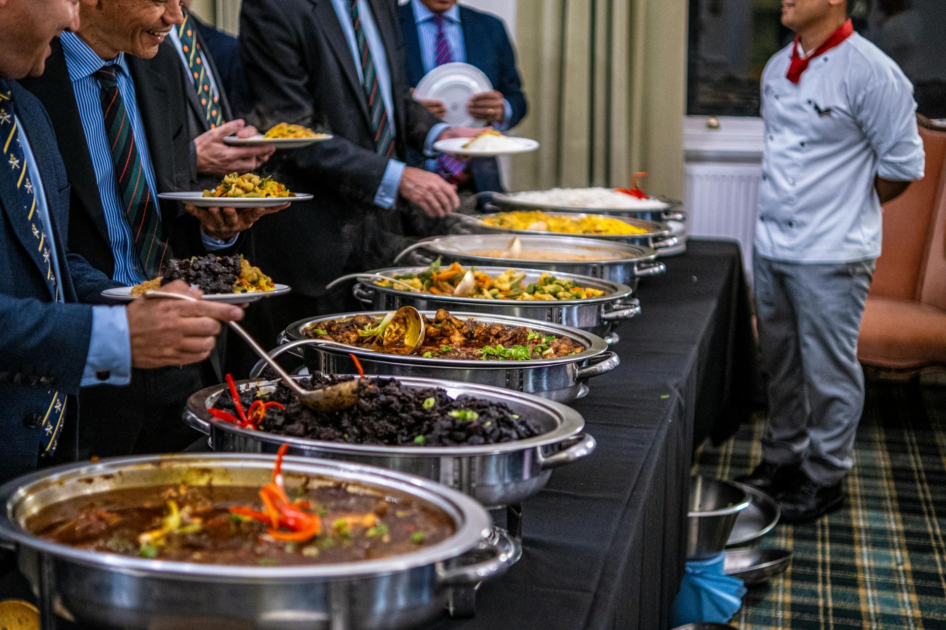 Attendees serving food from a buffet line with chafing dishes, overseen by a chef in uniform.