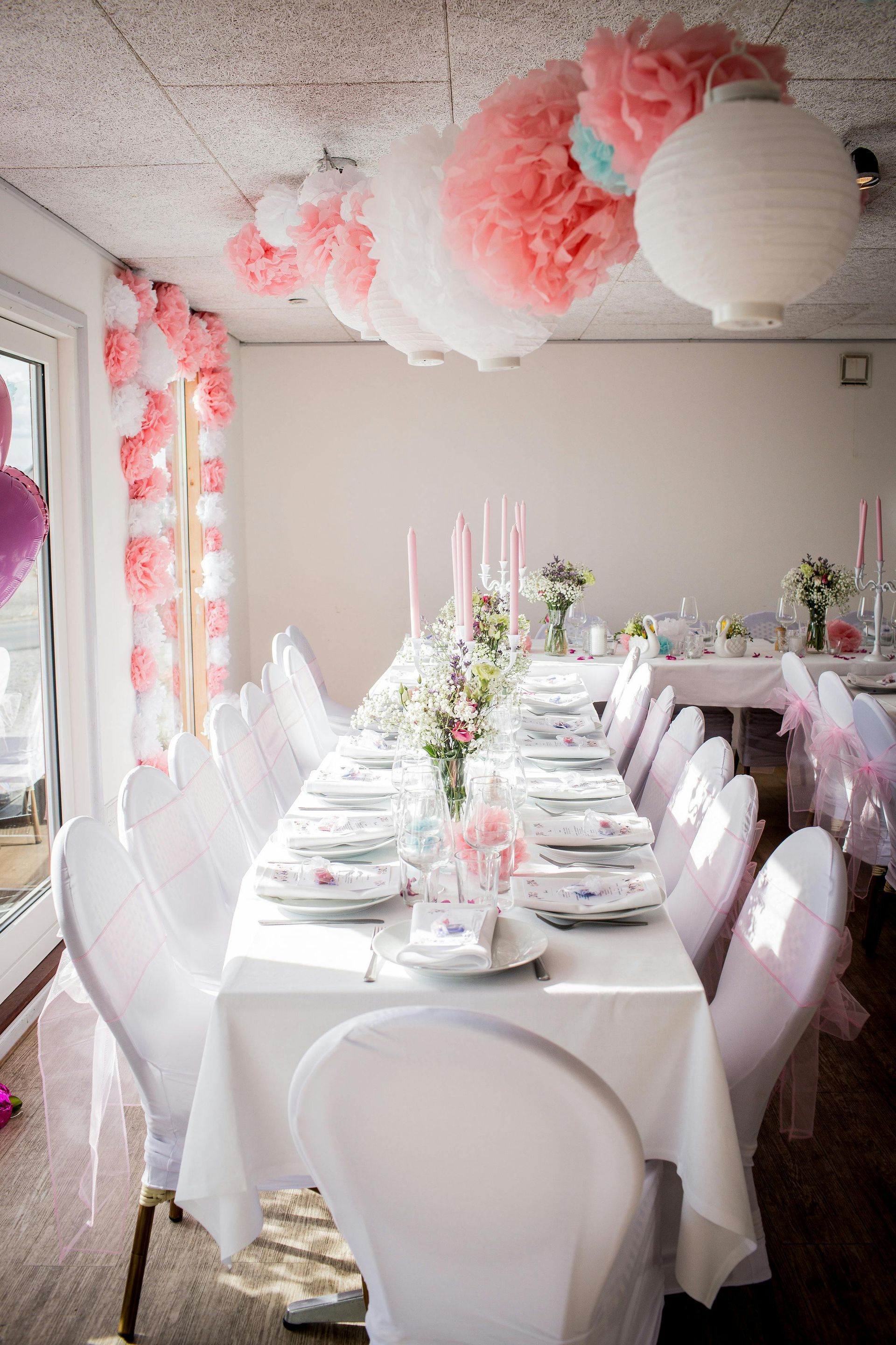 A long dining table set for a formal event, featuring white covers, pink decor, and hanging paper lanterns.