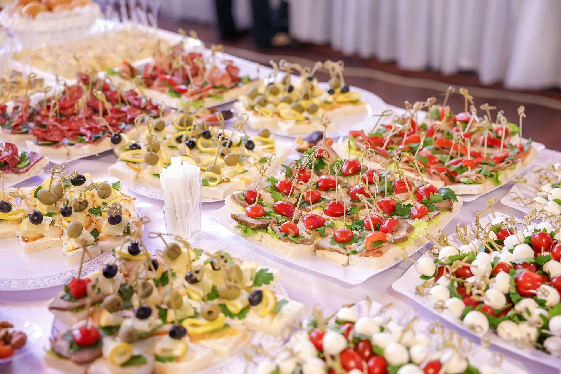 A spread of assorted party canapés and skewers on white platters, featuring tomatoes, olives, cheese, and meats.