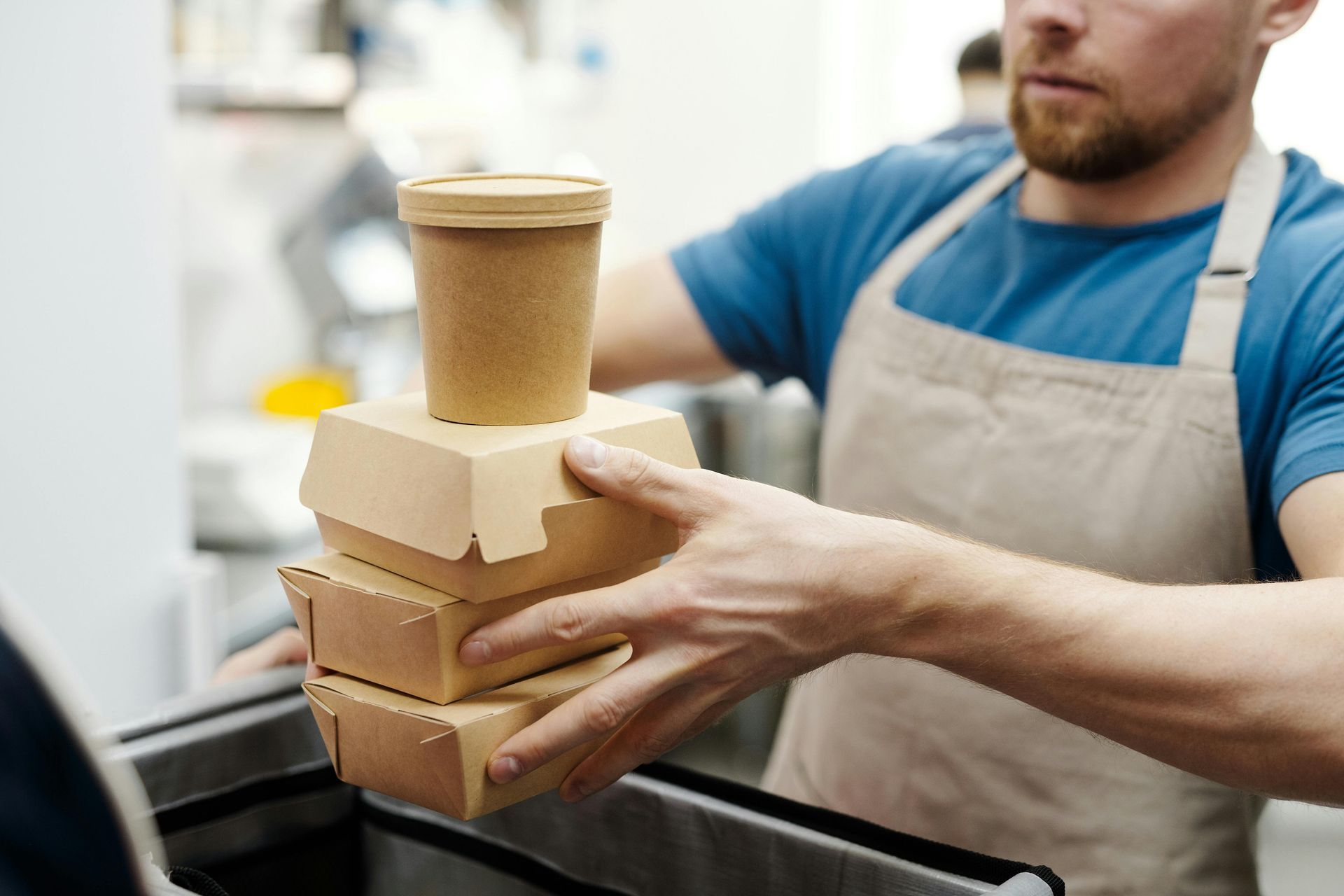 A person in a blue shirt and beige apron carefully places a stack of cardboard food containers into a thermal bag.