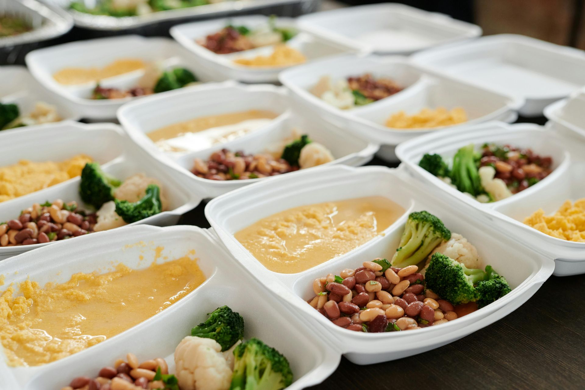 Multiple white to-go containers arranged on a surface, each holding scrambled eggs, beans, broccoli, and cauliflower.