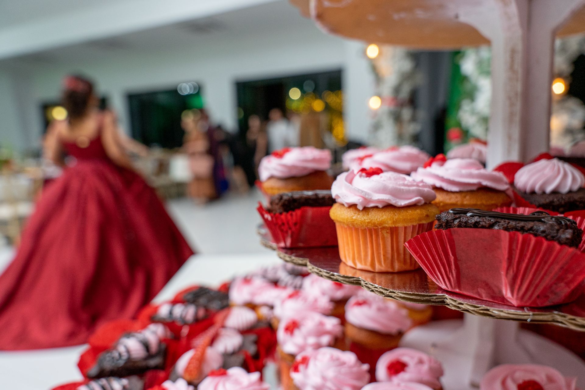 Cupcakes with pink frosting on a tiered stand, with a blurred person in a red formal gown in the background.