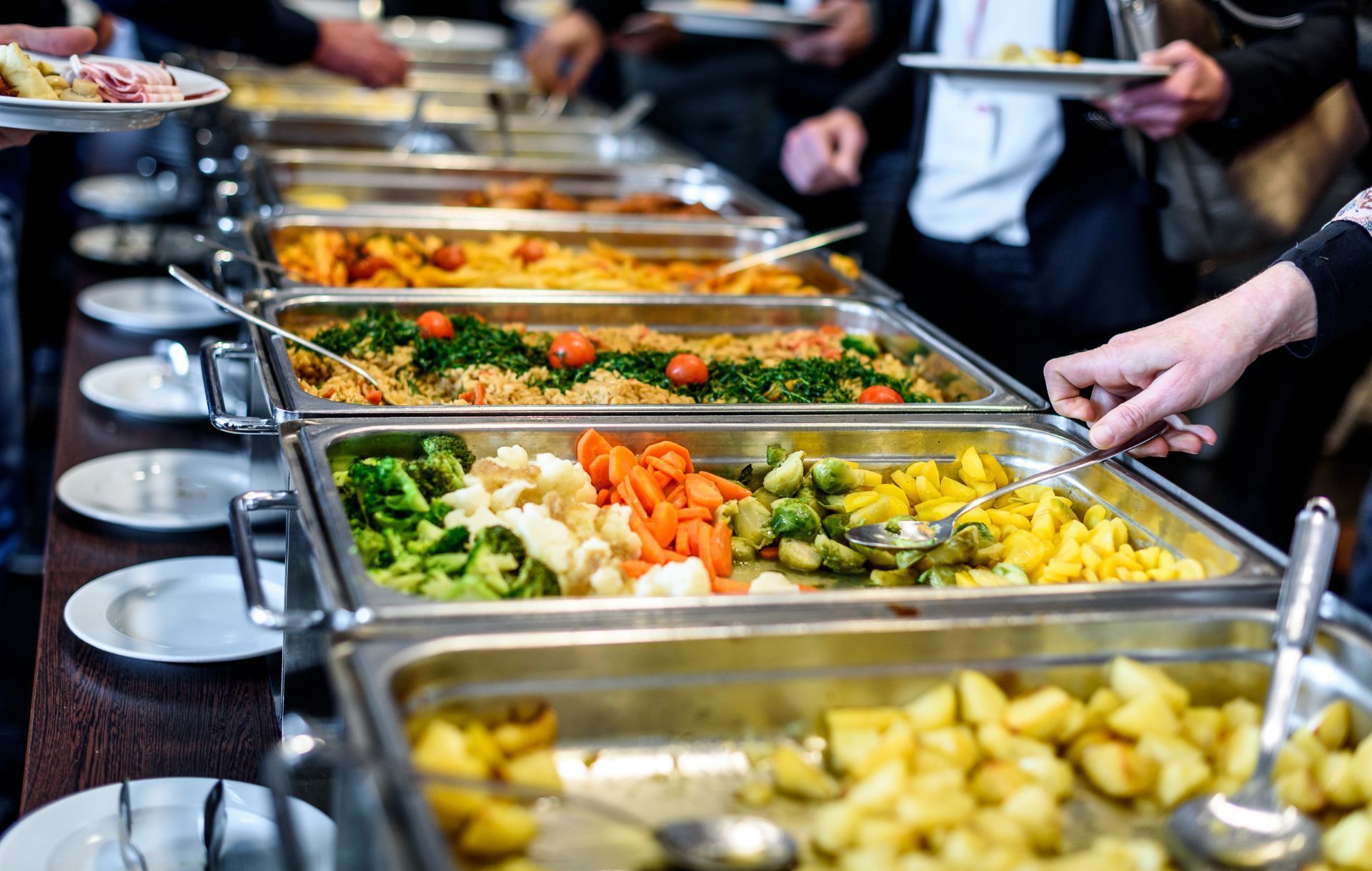 People serve themselves at a buffet with multiple metal warming trays filled with colorful vegetables and potatoes.