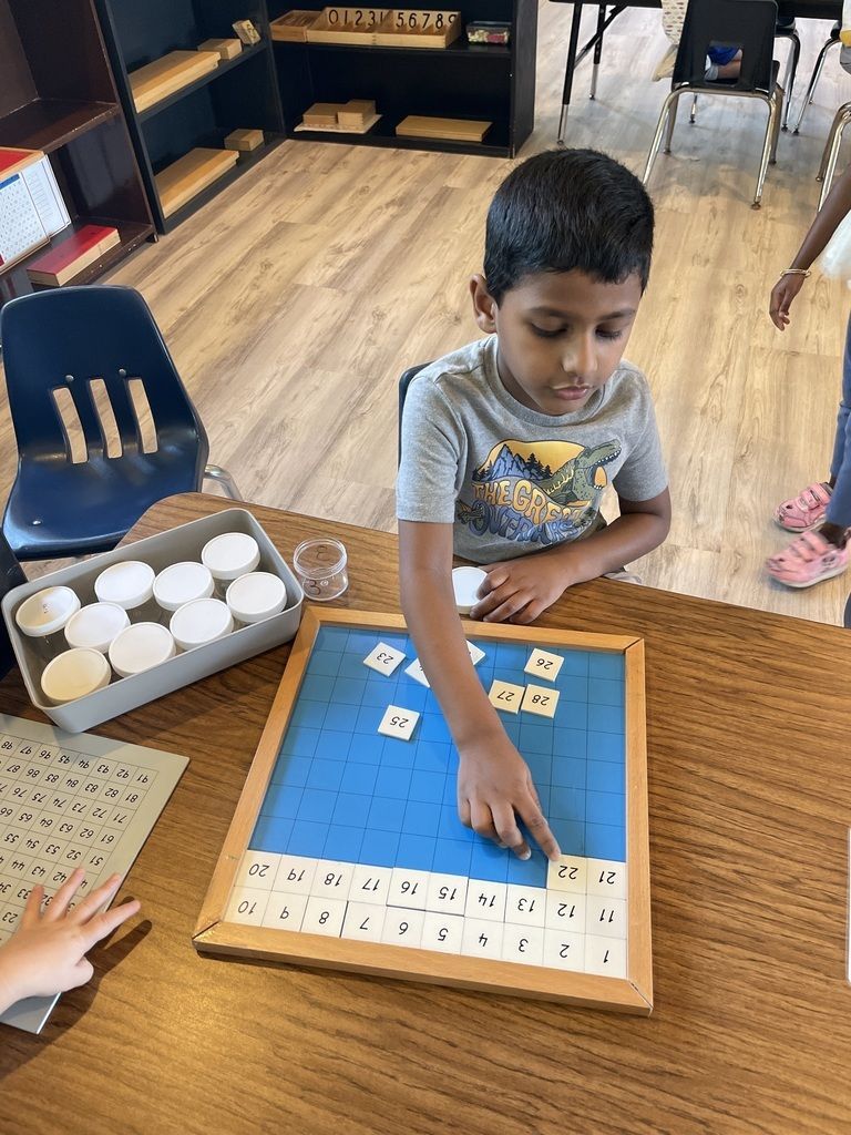 Child placing a tile on a blue board game at a classroom table with letter and number pieces nearby