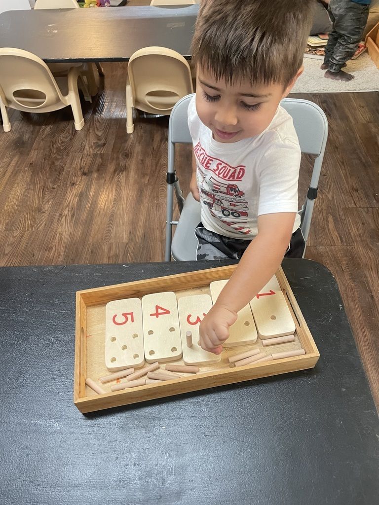 Boy playing with a wooden shape puzzle at a table in a classroom.
