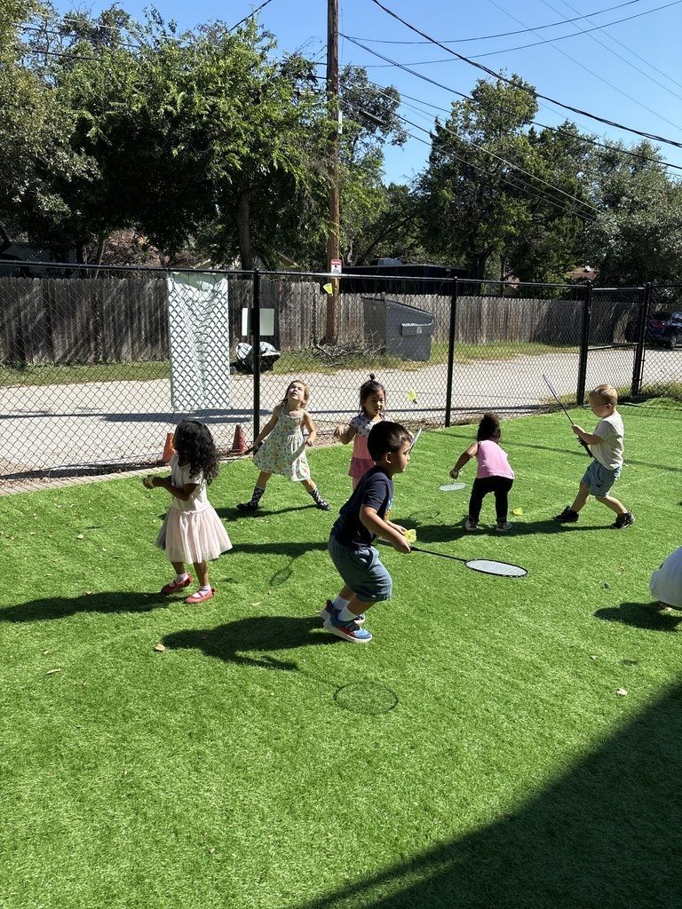 Children playing on green turf in a sunny fenced playground with trees in the background