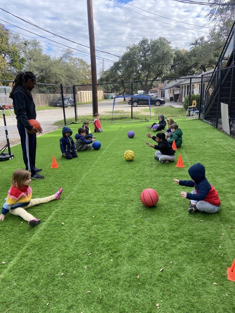 Children sit in a grassy fenced field during an outdoor soccer activity with colorful balls and cones.