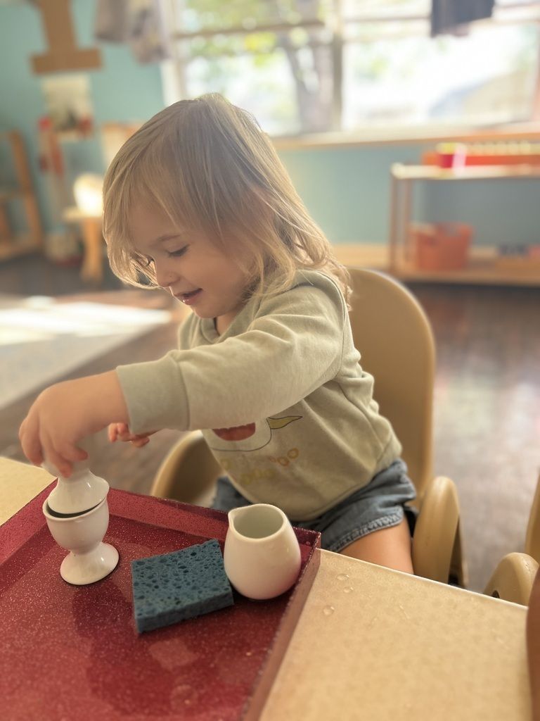 Child playing with stacking toys at a red table in a sunlit classroom