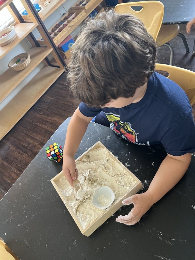 Child playing with sand at a classroom table, using a tray and a small white cup.