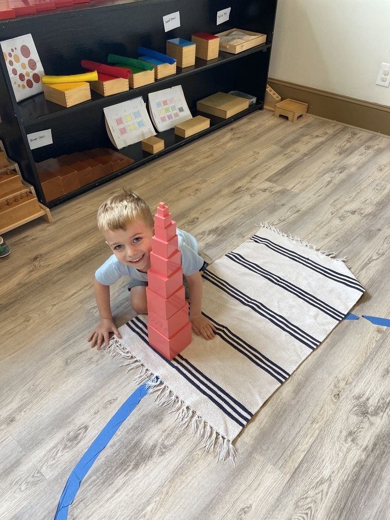 Child on a classroom rug beside a tall pink block tower and striped mat, with shelves of toys in the background