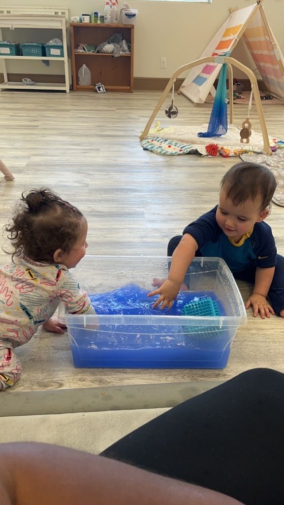Two toddlers play with water and a blue sensory bin in a classroom, with a teepee in the background.