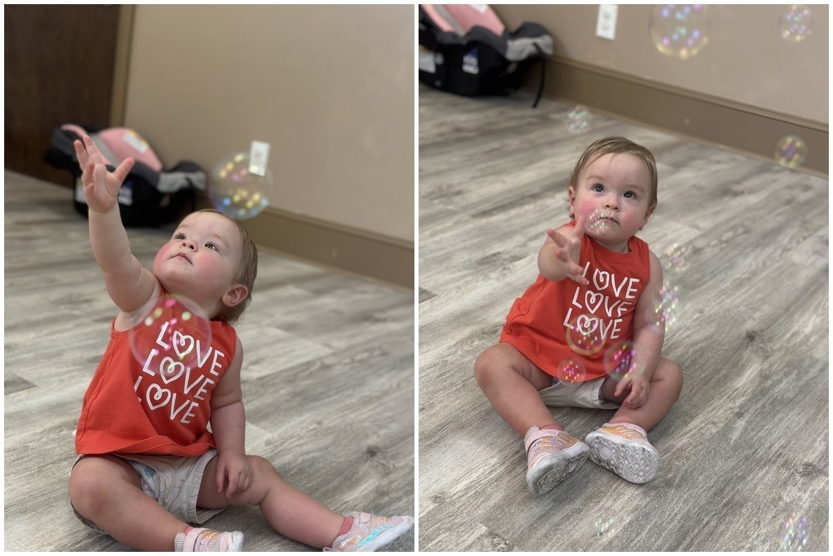 Child in red shirt sitting on the floor and reaching up to pop bubbles in a playroom.