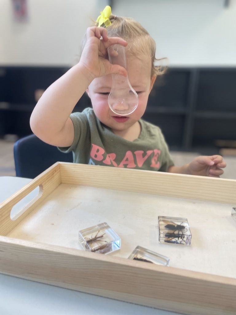 Child blowing through a tube over a wooden tray with small objects inside, in a classroom setting