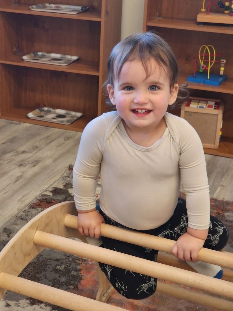 Smiling toddler standing in a wooden play area, holding a ladder rail and looking at the camera.