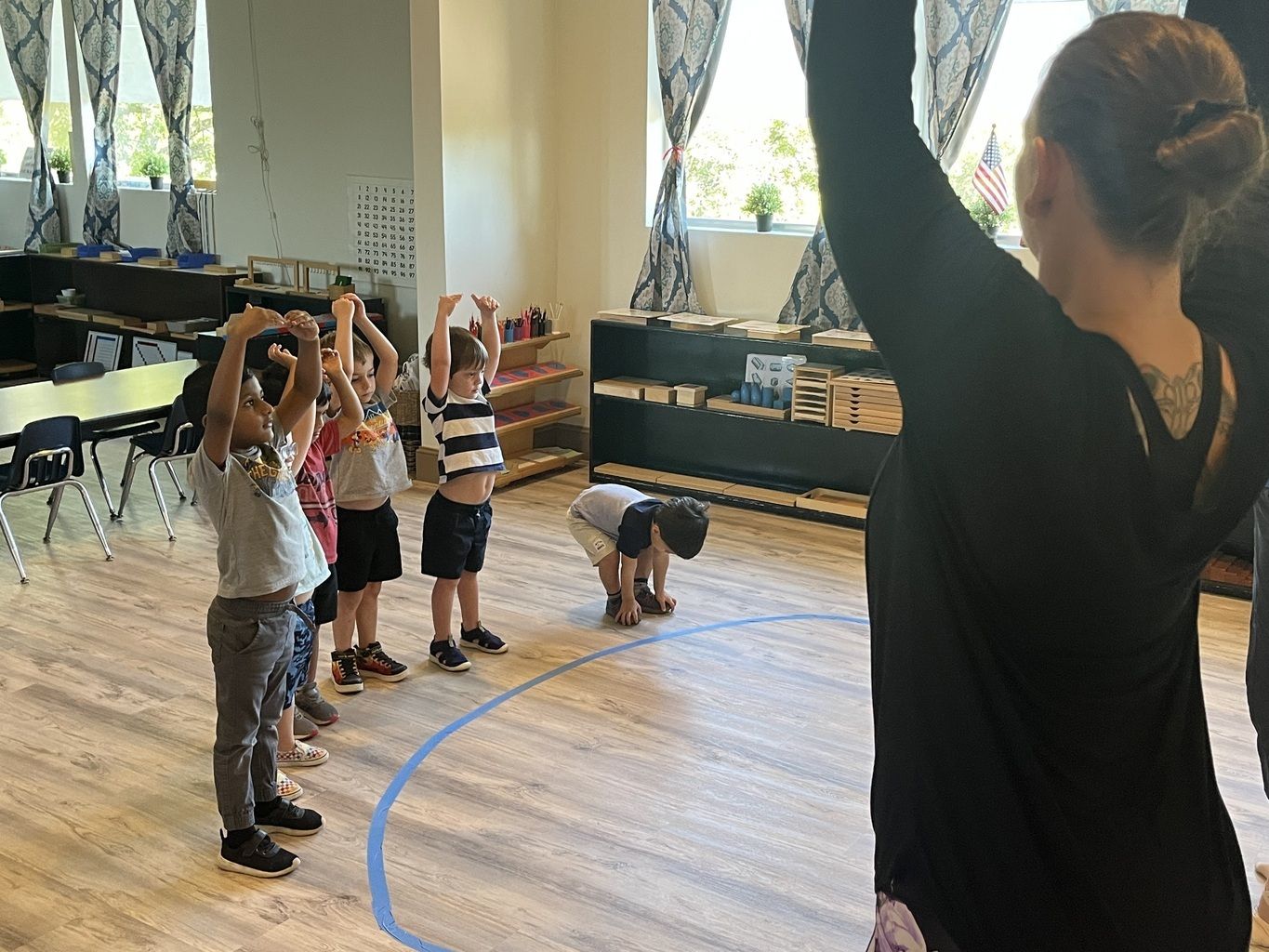 Children stretching in a gym while a teacher demonstrates overhead arm raises