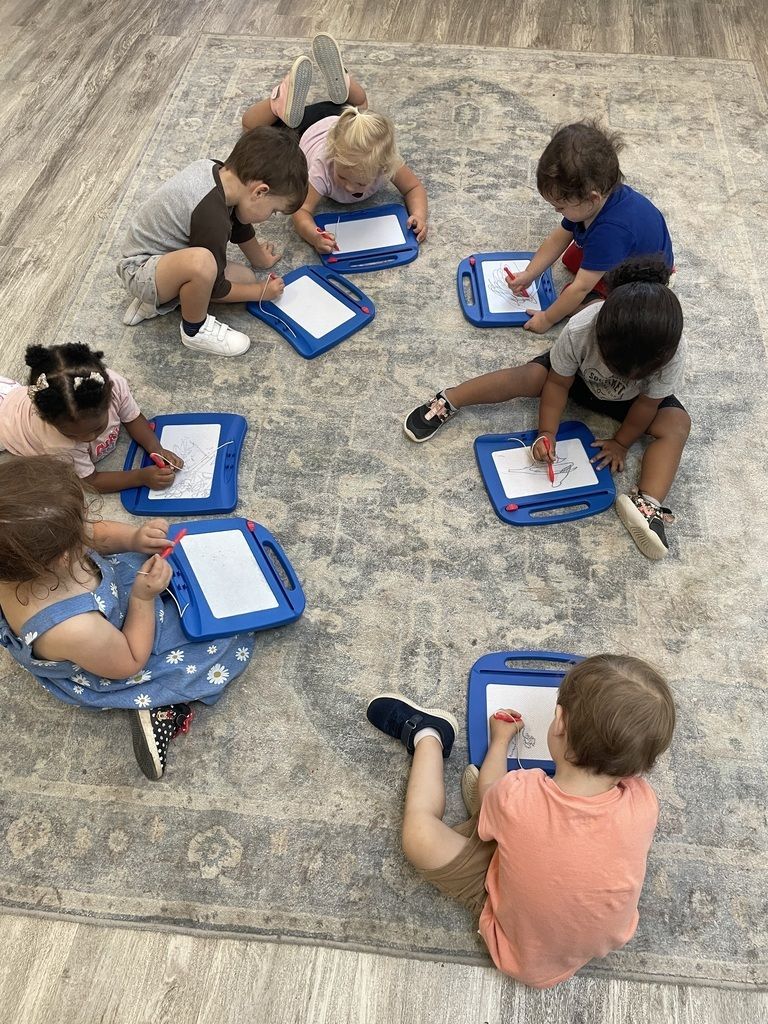 Children sitting in a circle on a rug, writing on blue clipboards with markers.