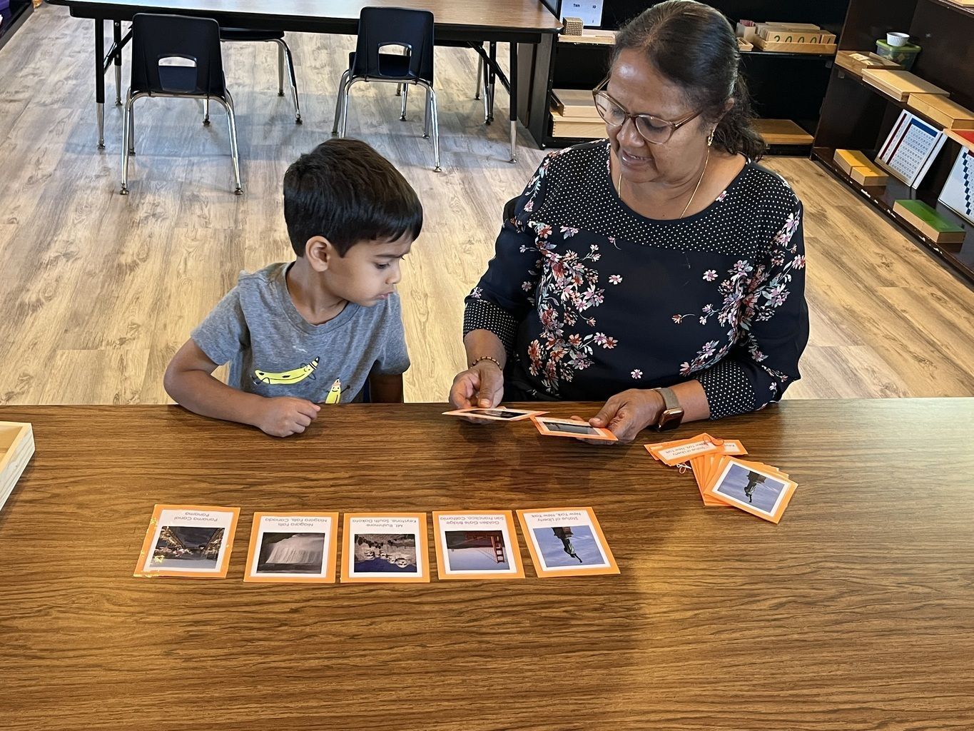 Adult helping a child arrange photo cards on a wooden table in a classroom