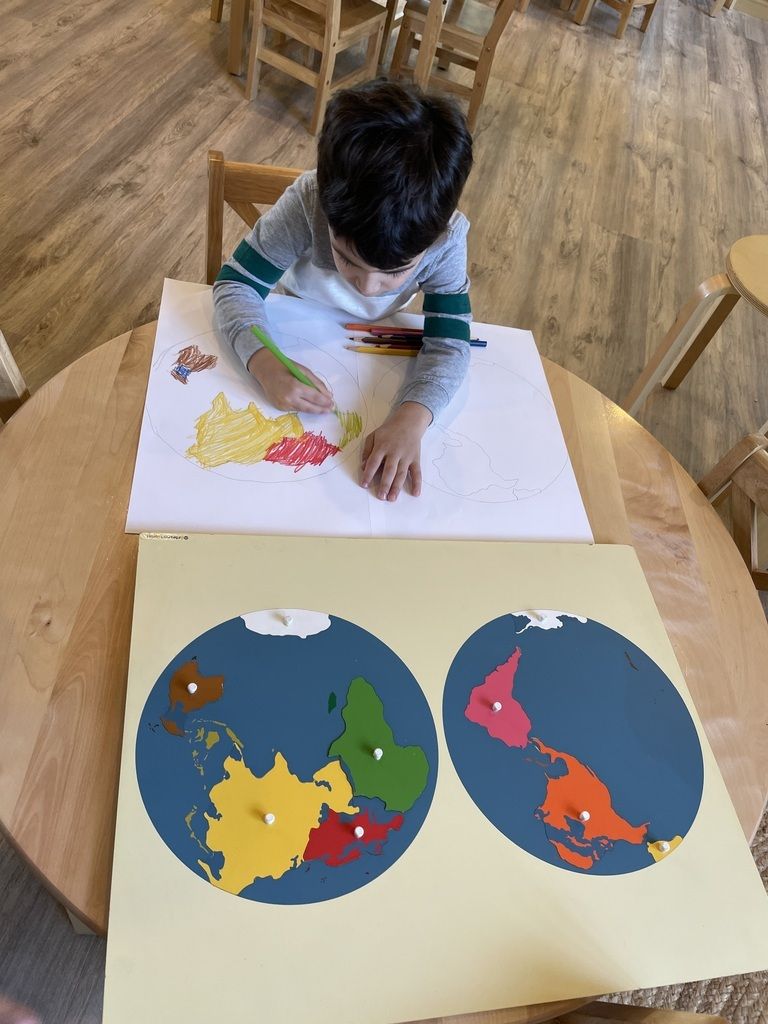 Child coloring a world map at a table, with two completed globe drawings in blue circles below.