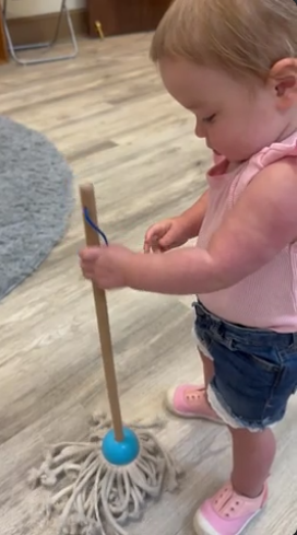 Toddler mopping a wood floor with a white mop and blue handle, wearing a pink shirt and jeans