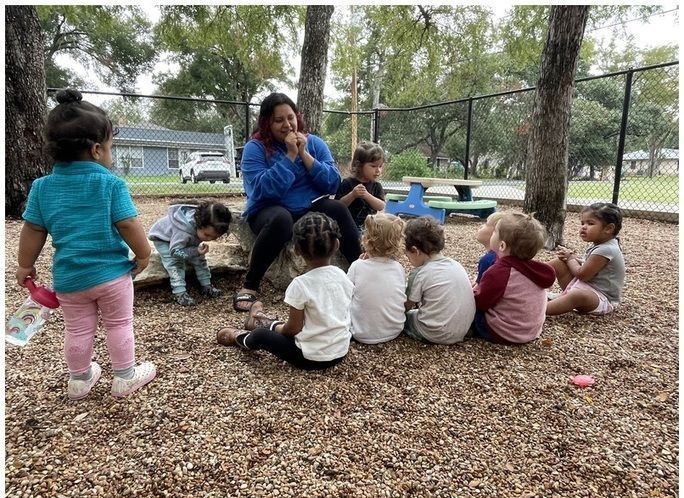 Children sit with a teacher on wood chips in a fenced playground, listening together outdoors.