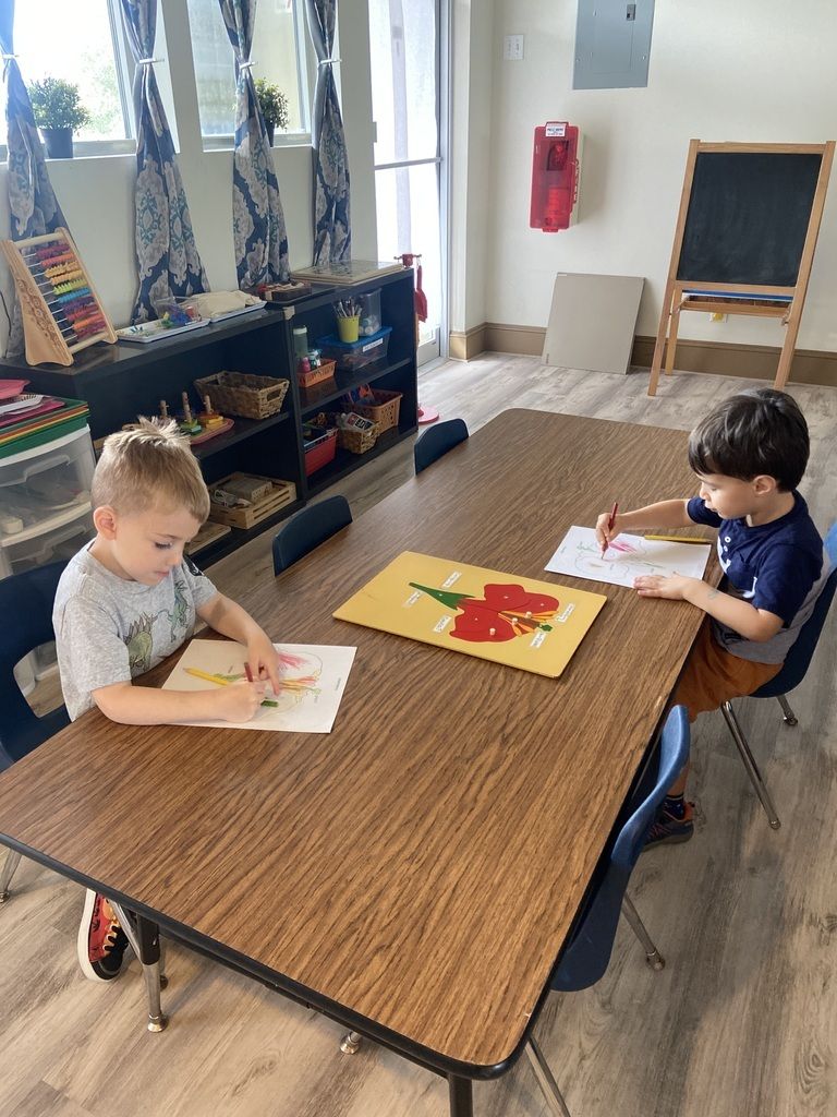 Two children coloring at a table in a classroom, with bookshelves and a chalkboard nearby