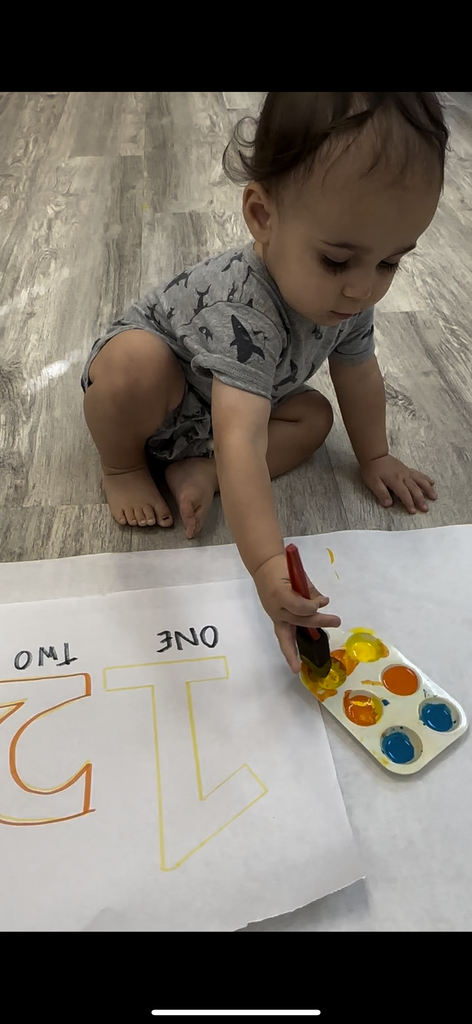Toddler painting numbers on paper with watercolor set on a floor.