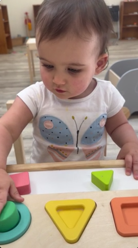 Toddler playing with a shape sorter, pressing a green button and looking down at the colorful blocks.