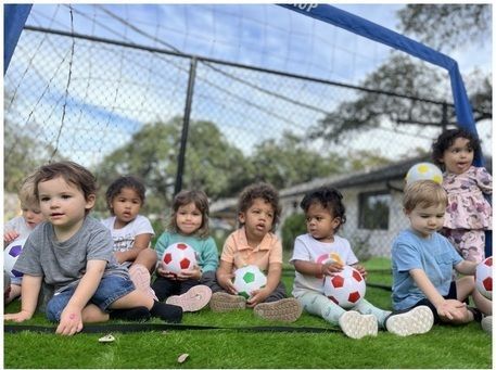 Children sitting on grass in front of a soccer goal, with colorful balls around them.