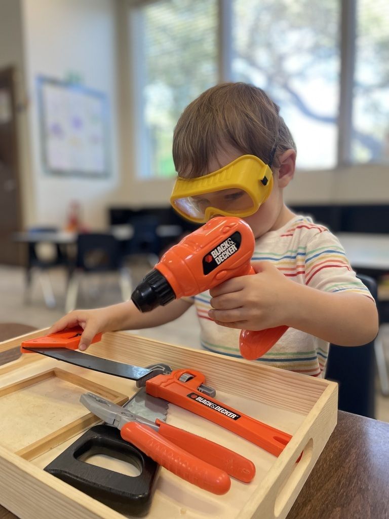 Child wearing safety goggles using an orange toy drill at a woodworking table with tools.