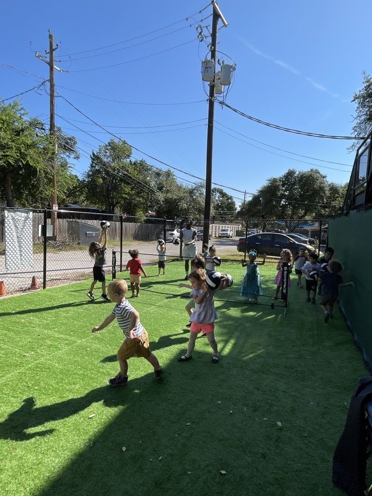 Children playing on green turf in an outdoor fenced playground on a sunny day.
