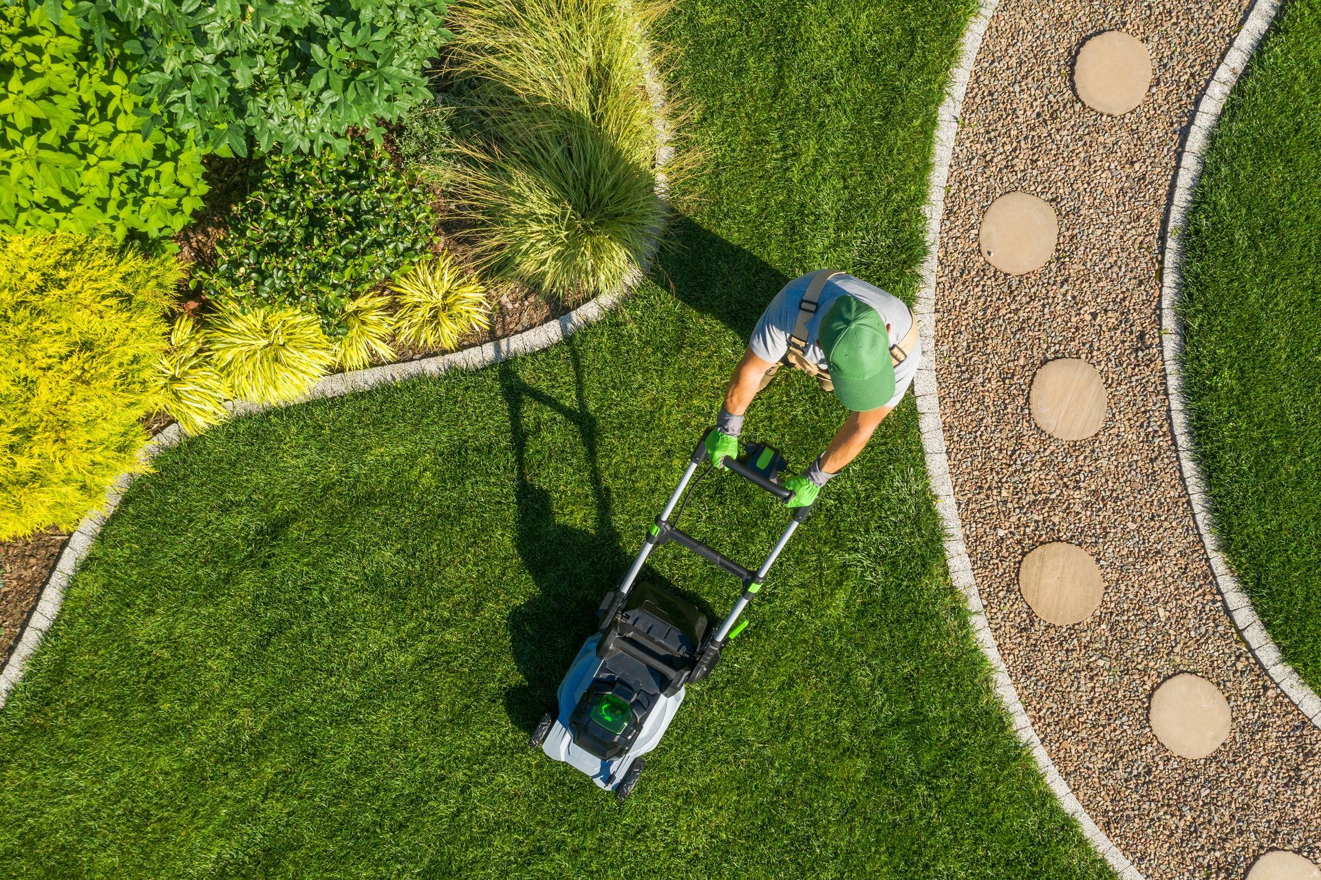 Person mowing a green lawn with a black and gray lawnmower next to a garden path.