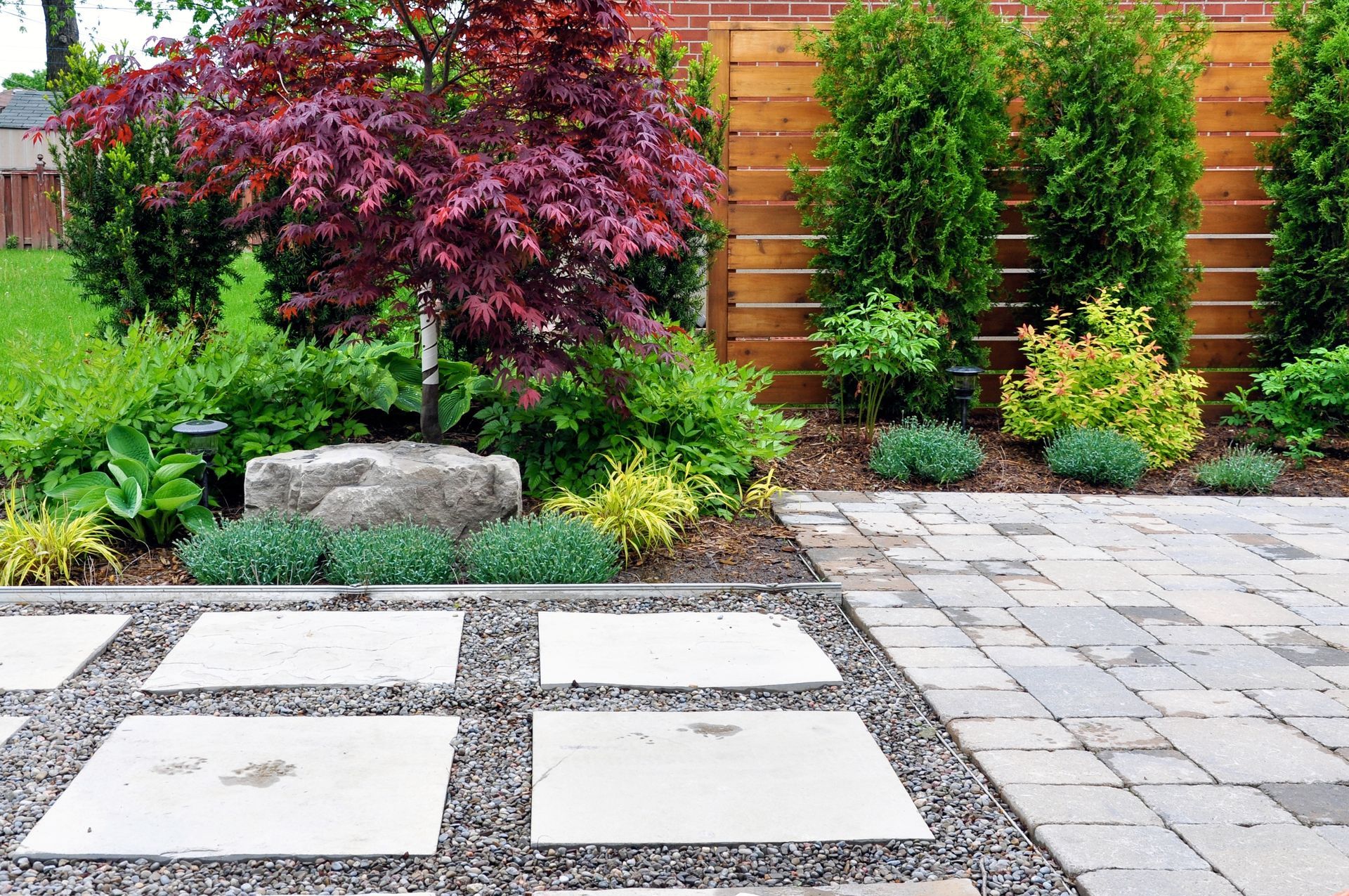 Landscaped backyard with stone patio, gravel, and diverse green and red foliage near a wooden fence.