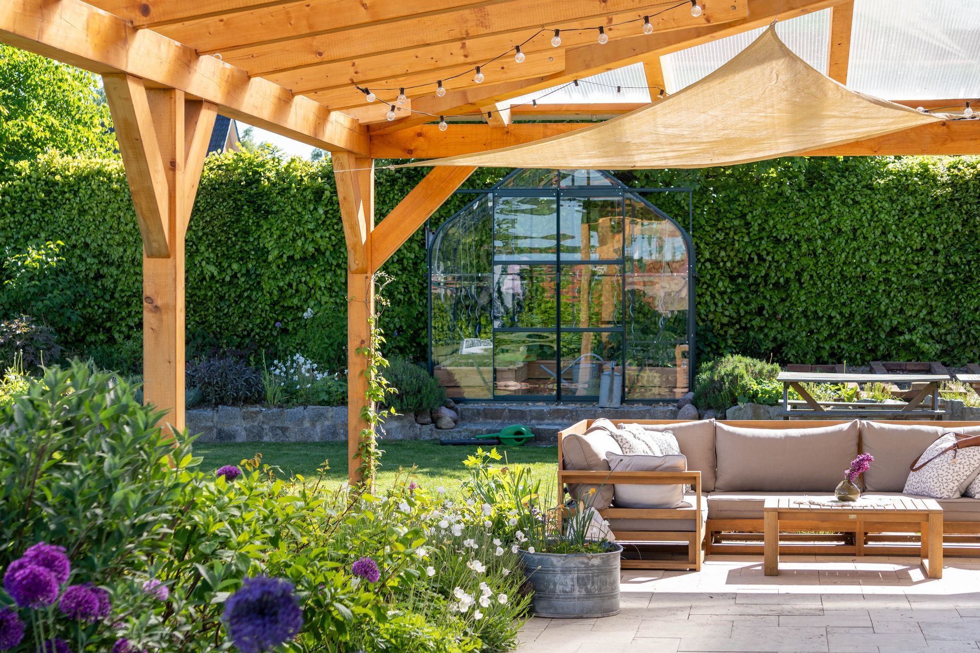 Wooden pergola over a patio with outdoor seating and a greenhouse in a garden